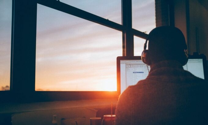 Man mit Kopfhörern der vor einem PC-Bildschirm sitzt mit einem Sonnenuntergang im Hintergrund